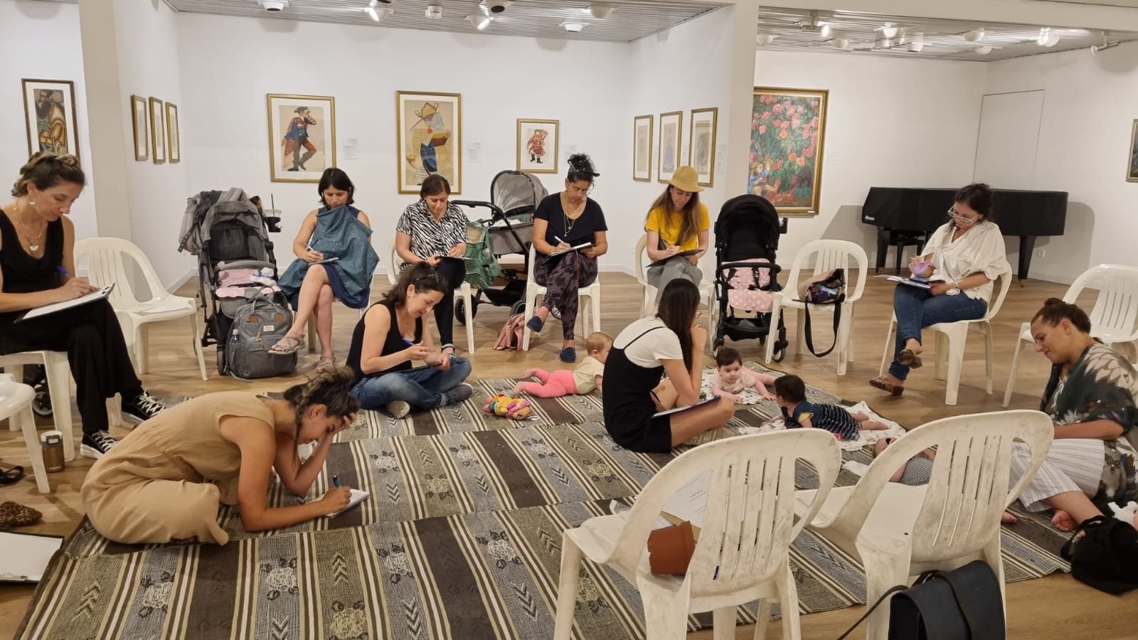 אמהות כותבות בסדנה, חלקן עם תינוקות על השטיח, בחדר גלריה